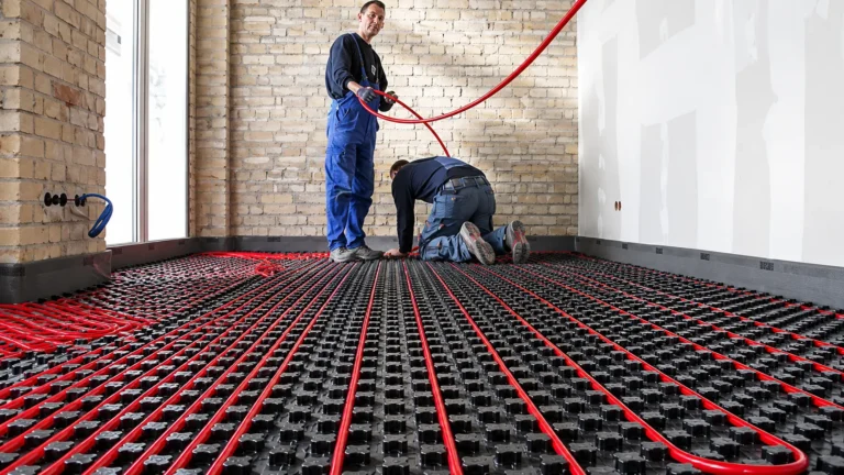 Two installers laying red hydronic tubing into black castellated panels across a subfloor before the finished floor goes down