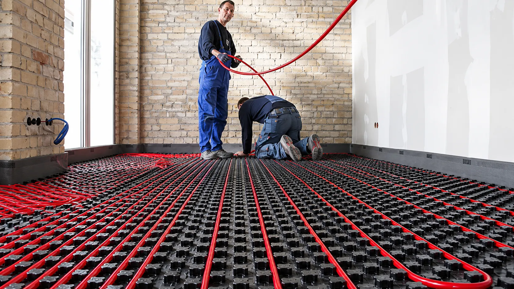 Two installers laying red hydronic tubing into black castellated panels across a subfloor before the finished floor goes down