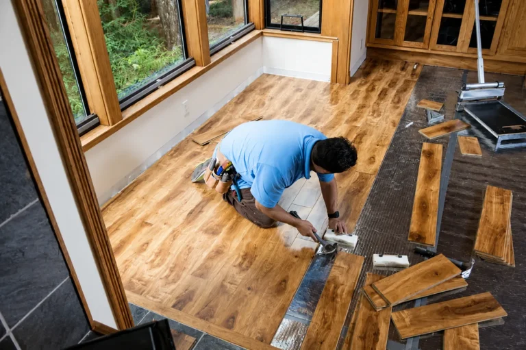 Installer using a tapping block on golden plank flooring over silver underlay in a room with wood-framed windows