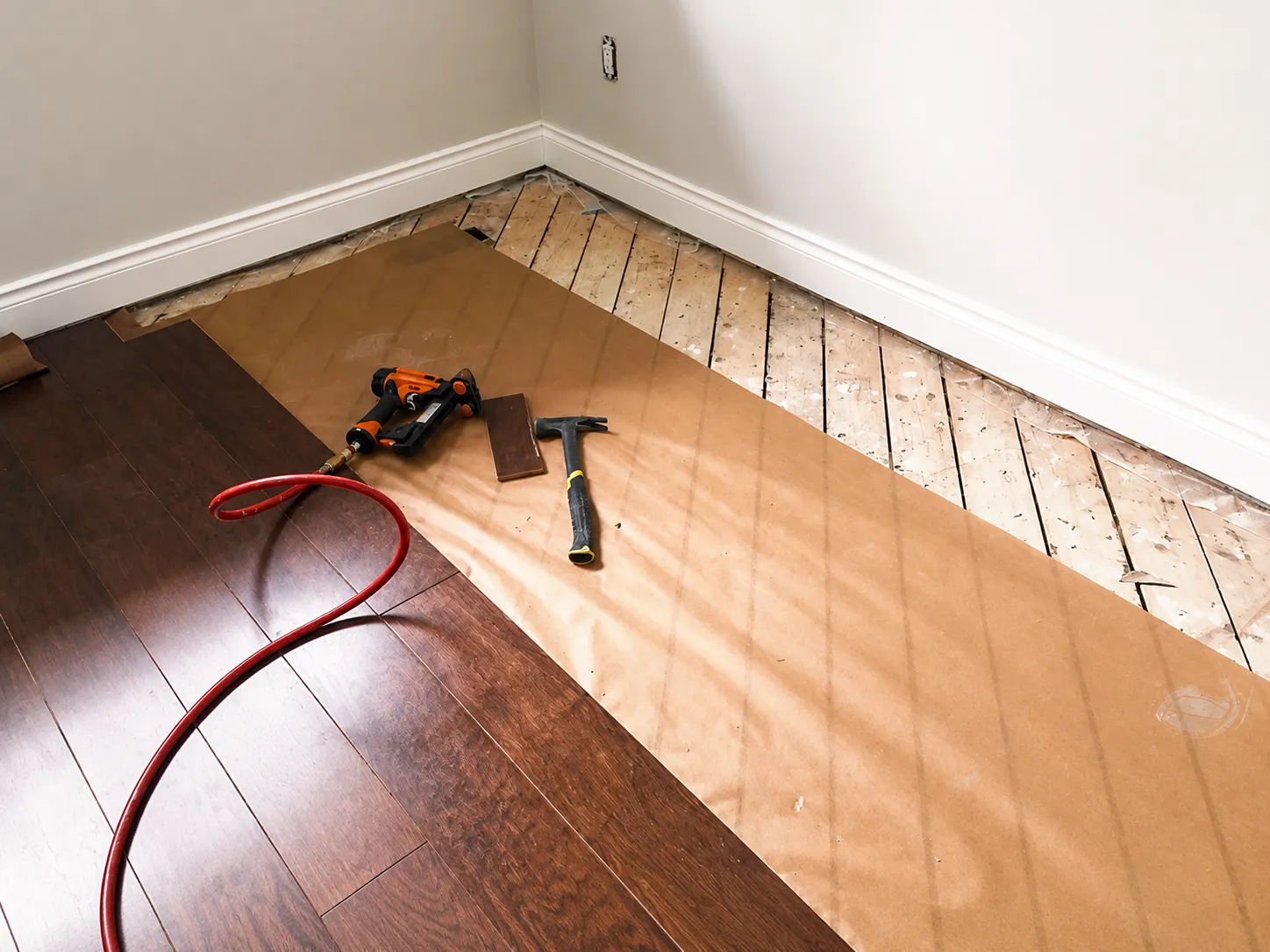 Kraft paper underlay rolled over an old plank subfloor beside finished dark hardwood, with a flooring nailer and hammer on top